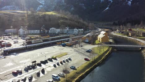 Flamsbanen-railway-station-seen-during-morning-sunrise---Aerial-from-above-Flam-river-looking-at-train-waiting-on-station-before-departure---Norway