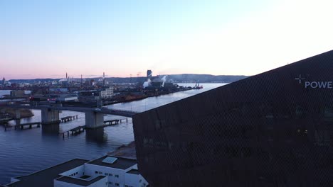 Flying-above-energy-plus-house-Powerhouse-in-Porsgrunn-Norway-during-late-evening---Logo-on-house-visible-and-city-bridge-with-channel-and-Heroya-industrial-area-in-Background