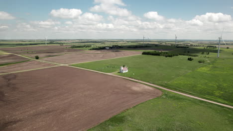 Aerial-approach-towards-small-church-in-sunny-field,-wind-turbines-turning-in-background