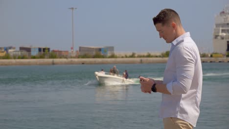 Ein-Junger-Kaukasischer-Mann-Im-Profil-Auf-Seinem-Smartphone-In-Einem-Hafen-In-Südfrankreich,-Während-Im-Hintergrund-Ein-Kleines-Boot-Mit-Menschen-An-Bord-Fährt