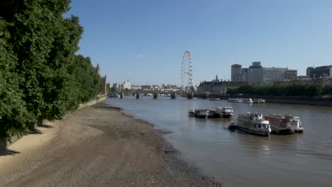 Establishing-shot-of-several-moored-boats-along-the-River-Thames-In-Low-Tide,-in-the-distance-the-iconic-London-Eye-Ferris-wheel-and-the-Westminster-Bridge-on-a-beautiful-sunny-day,-London,-England