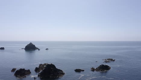 Ocean-aerial-flies-past-jagged-rocks-toward-flat-blue-water-horizon