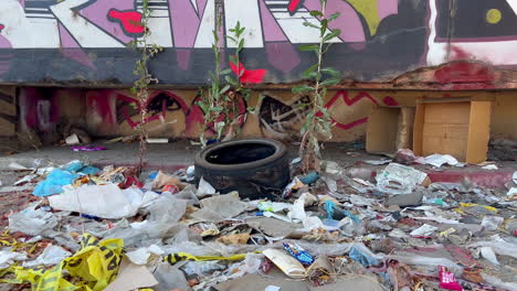 Basura-Esparcida-En-La-Acera-De-La-Ciudad-Durante-El-Día,-Neumáticos-Y-Montones-De-Envoltorios-Que-Rodean-La-Pared-De-Graffiti