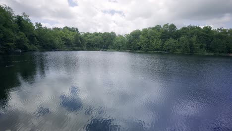 Cloudy-Sky-Reflecting-On-Calm-Lake-With-Lush-Vegetation-In-Background
