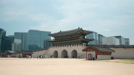Tourists-walking-through-Gwanghwamun-gate-to-the-square-of-Gyeongbokgung-Palace-with-Seoul's-business-high-rise-office-buildings-against-blue-sky---distant-view