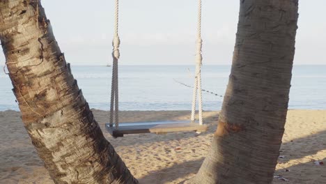 A-close-up-shot-of-a-swaying-swing-suspended-between-the-trunks-of-two-coconut-trees,-the-view-overlooking-the-beautiful-ocean-waters-of-the-Gulf-of-Thailand-in-Pattaya,-Chon-Buri,-Thailand