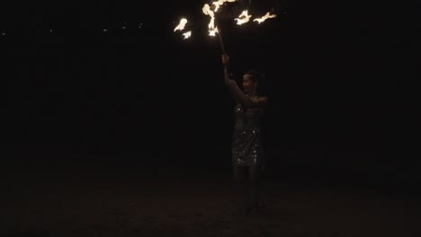 A-female-Thai-dancer-pirouetting-while-twirling-and-waving-a-fire-torch-during-a-dance-routine-entertaining-a-group-of-tourists-on-a-beach-in-Pattaya,-Chon-Buri,-Pattaya