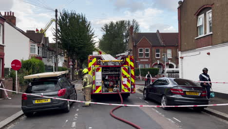A-London-Fire-Brigade-pump-firetruck-and-turntable-aerial-ladder-firetruck-are-behind-a-cordon-tape-at-the-scene-of-a-house-on-a-residential-road