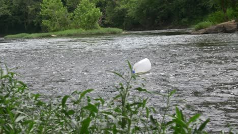 milk-jug-floats-down-river-slowly-behind-foliage-towards-old-concrete-bridge-on-a-cloudy-day-slow-motion
