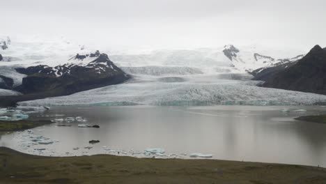 Island-Gletscher-Mit-Wasser,-Grünem-Gras,-Blauem-Eis-Und-Nebel-Mit-Nach-Oben-Bewegendem-Drohnenvideo