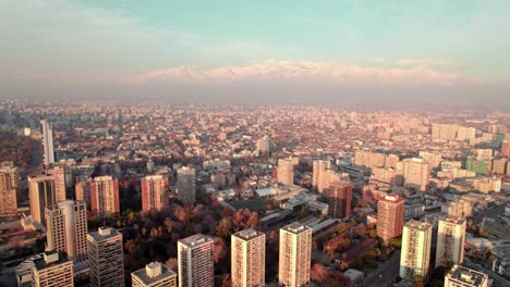 Aerial-dolly-in-of-Downtown-Santiago-city-buildigns-and-snowcapped-Andean-mountain-range-in-the-background-at-sunset,-Chile
