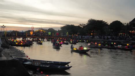 Festival-De-Los-Faroles-Hoi-An-Vietnam,-Vista-De-La-Puesta-De-Sol-De-La-Tarde-Del-Lapso-De-Tiempo-Del-Tráfico-De-Canoas-De-Barcos-Iluminados-En-El-Canal-Del-Río,-Pasando-Por-Debajo-Del-Puente,-Turistas-A-Bordo,-Multitud-De-Visitantes-En-Segundo-Plano