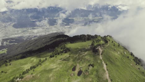 Aerial-drone-shot-of-a-grass-covered-mountain-top-with-a-small-trail-leading-up-with-clouds-around