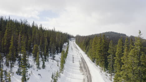 Low-aerial-reveal-backward-of-sunny-and-snow-covered-back-roads-in-the-Colorado-mountains-surrounded-by-bright-green-pine-tree-forests-with-blue-skies