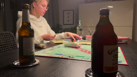 Mujer-Caucásica-Clasificando-Cartas-De-Juego-De-Monopolio-Con-Botellas-De-Cerveza-En-La-Mesa