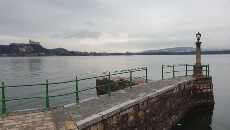 Seagulls-on-green-handrail-of-Arona-pier-on-Maggiore-lake-with-Angera-fortress-in-background