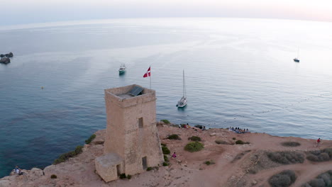 Stone-tower-with-maltese-flag-on-a-cliff-above-a-bay-with-boats,aerial