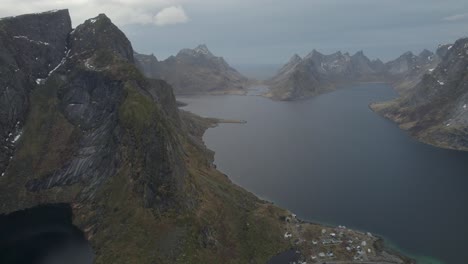 Aerial-view-overlooking-a-boat-driving-on-a-mountainous-fjord,-dark,-gloomy-fall-day-in-Lofoten,-Norway---pan,-drone-shot