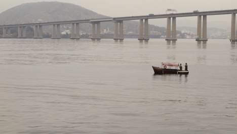 Two-Local-Men-Stand-On-Small-Boat-Floating-At-Guanabara-Bay-Near-Rio-Niteroi-Bridge-In-Brazil