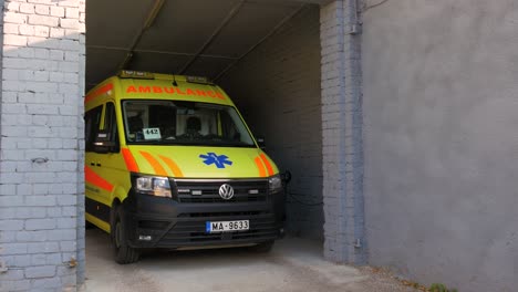 The-yellow-paramedic-ambulance-vehicle-van-parked-in-white-brick-garage,-sunny-day,-medium-shot