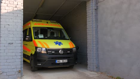 La-Furgoneta-De-Ambulancia-Paramédica-Amarilla-Estacionada-En-Garaje-De-Ladrillo-Blanco,-Día-Soleado,-Plano-Medio