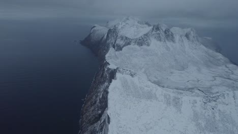 Aerial-view-overlooking-the-Hesten-mountain-in-gloomy,-snowy-Senja,-Norway---reverse,-tilt-drone-shot