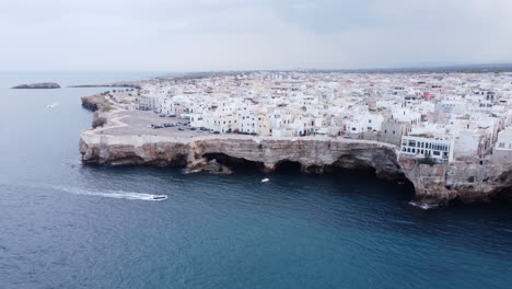 Luxury-motorboat-passes-by-the-Italian-village-of-Polignano-a-Mare,-aerial