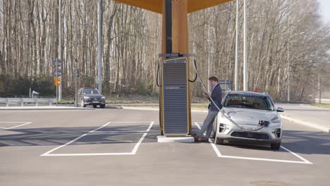 Man-In-Suit-Leaning-On-His-Car-Charging-At-The-Electric-Station-In-Leuven,-Belgium