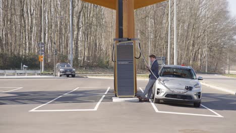 Hombre-De-Traje-Apoyado-En-Su-Auto-Cargándose-En-La-Estación-Eléctrica-En-Lovaina,-Bélgica