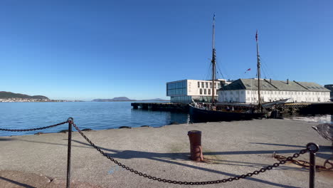 Old-wooden-ship-motors-out-of-Alesund-Harbor,-Norway-on-a-beautiful-clear-calm-day-with-a-brilliant-blue-sky-and-calm-seas,-panning-right-to-left