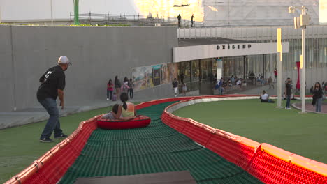 man-pulling-tubby-with-kids-going-down-tubby-rim-to-slide-on-green-grass-downhill,-outdoors