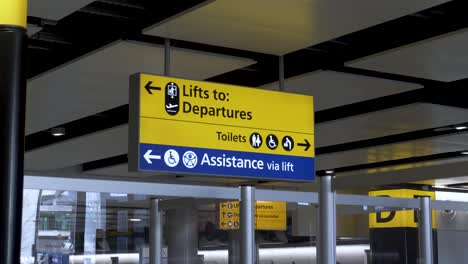 Señalización-Direccional-Amarilla-Y-Azul-En-La-Terminal-3-Del-Aeropuerto-Internacional-De-Heathrow,-La-Señal-Proporciona-A-Los-Pasajeros-Una-Guía-Para-Llegar-A-La-Terminal-De-Salida,-Aseos-Y-Ascensor-De-Pasajeros-Asistido,-Londres,-Reino-Unido