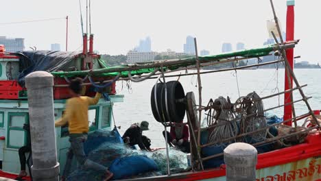 Un-Pescador-Se-Levanta-Para-Ir-Al-Costado-Del-Bote-Mientras-Otros-Reparan-Redes,-Muelle-De-Pesca-De-Pattaya,-Chonburi,-Tailandia