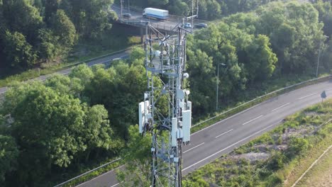 Antena-De-Torre-De-Comunicación-5g-En-El-Campo-Británico-Con-Vehículos-Que-Viajan-En-La-Vista-Izquierda-De-La-órbita-Aérea-De-Fondo-De-La-Carretera