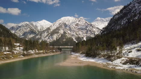 Idyllic-forest-banks-around-Plansee-lake-aerial-view-towards-snow-covered-Austria-mountains-landscape