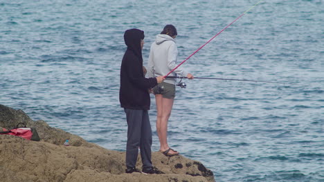 Little-Boy-Watching-Adults-Fishing-On-The-Sea-With-A-Fishing-Rod-In-Porthallow,-Cornwall,-UK