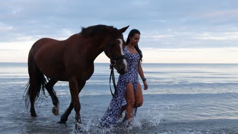 A-beautiful-girl-with-long-hair-in-a-blue-dress-walks-her-horse-through-the-water-during-the-evening-in-Donabate,-Ireland