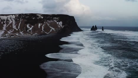Una-Hermosa-Toma-De-Drones-De-La-Playa-Negra-En-Islandia-Muestra-Las-Altas-Olas-Del-Mar-Y-El-Clima-Nublado-Mientras-La-Nieve-Cubre-Parte-De-La-Arena-Negra