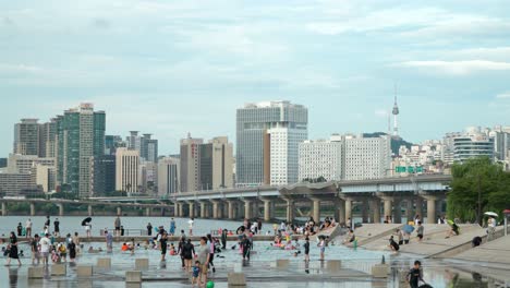 Yeouido-Hangang-Park-Cascade-Plaza-with-people-cooling-down-in-shallow-water-pools,-view-of-Mapo-bridge-and-urban-Seoul-landscape