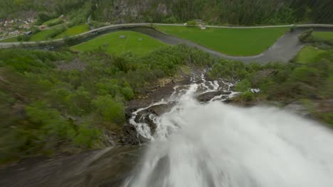 Drone-flight-down-the-splashing-waterfall-to-valley-with-road-and-cars-in-Norway