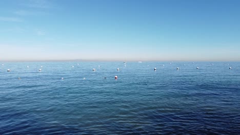 Drone-flies-from-Descando-Beach,-Catalina-Island,-California-out-to-the-ocean-revealing-buoys-and-boats-from-distant