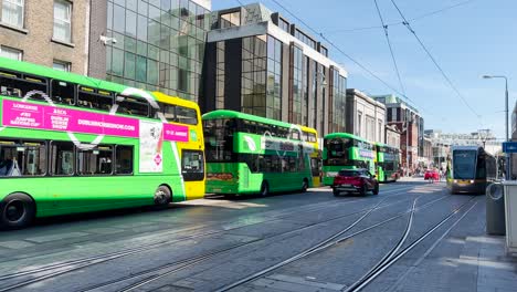 The-Dublin-tram-arrives-at-the-Abbey-Street-stop,-with-many-people-and-Dublin-buses-parked-along-the-street-on-a-sunny-day