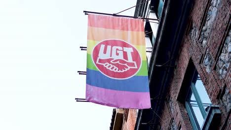 Cámara-Lenta-De-Una-Bandera-Del-Arco-Iris-Lgtbq-Ondeando-Apoyada-Por-La-Unión-Ugt-En-Un-Edificio-De-Ladrillo-En-Madrid,-España