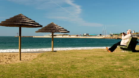 Old-man-sitting-at-beach-enjoys-lovely-weather,-taking-pictures-with-phone