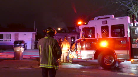 A-group-of-firefighters-gather-outside-of-a-burning-house,-Night,-Canada