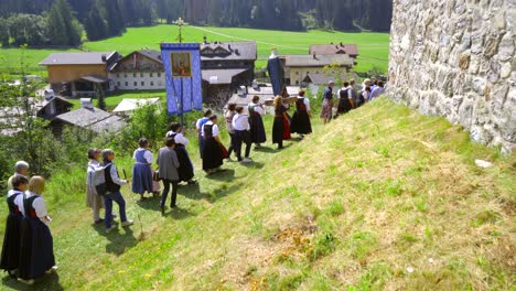 Worshippers-in-traditional-Tyrolean-costume-are-walking-on-grass-during-a-religious-procession-in-a-small-village-in-Tirol,-carying-a-blue-banner-and-a-statue-of-the-Virgin-Mary