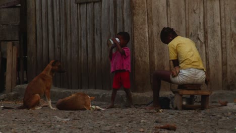 Eine-Afrikanische-Mutter-Rührt-Den-Topf-Draußen-Vor-Dem-Haus-Auf-Einer-Bank,-Wo-Ein-Kind-Trinkt-Und-Die-Tiere-Frei-Herumlaufen---Nahaufnahme