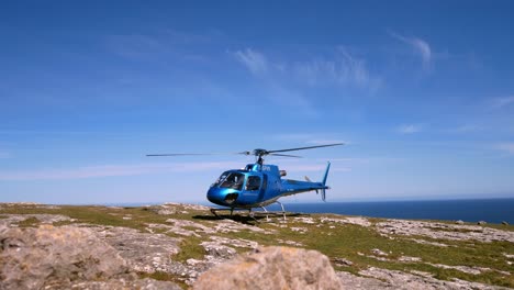 Private-helicopter-perched-on-mountain-cliff-summit-overlooking-blue-ocean-landscape-with-propeller-spin-right-dolly