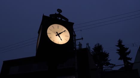 Silhouette-of-an-old-Illuminated-grandfather-clock-on-a-dark-evening-amongst-buildings-and-electrical-wires-trees-sky