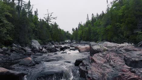 Drone-footage-flying-low-slowly-over-a-rushing-river-with-rapids-surrounded-by-forest-with-boulders-and-rocks-on-a-cloudy-day